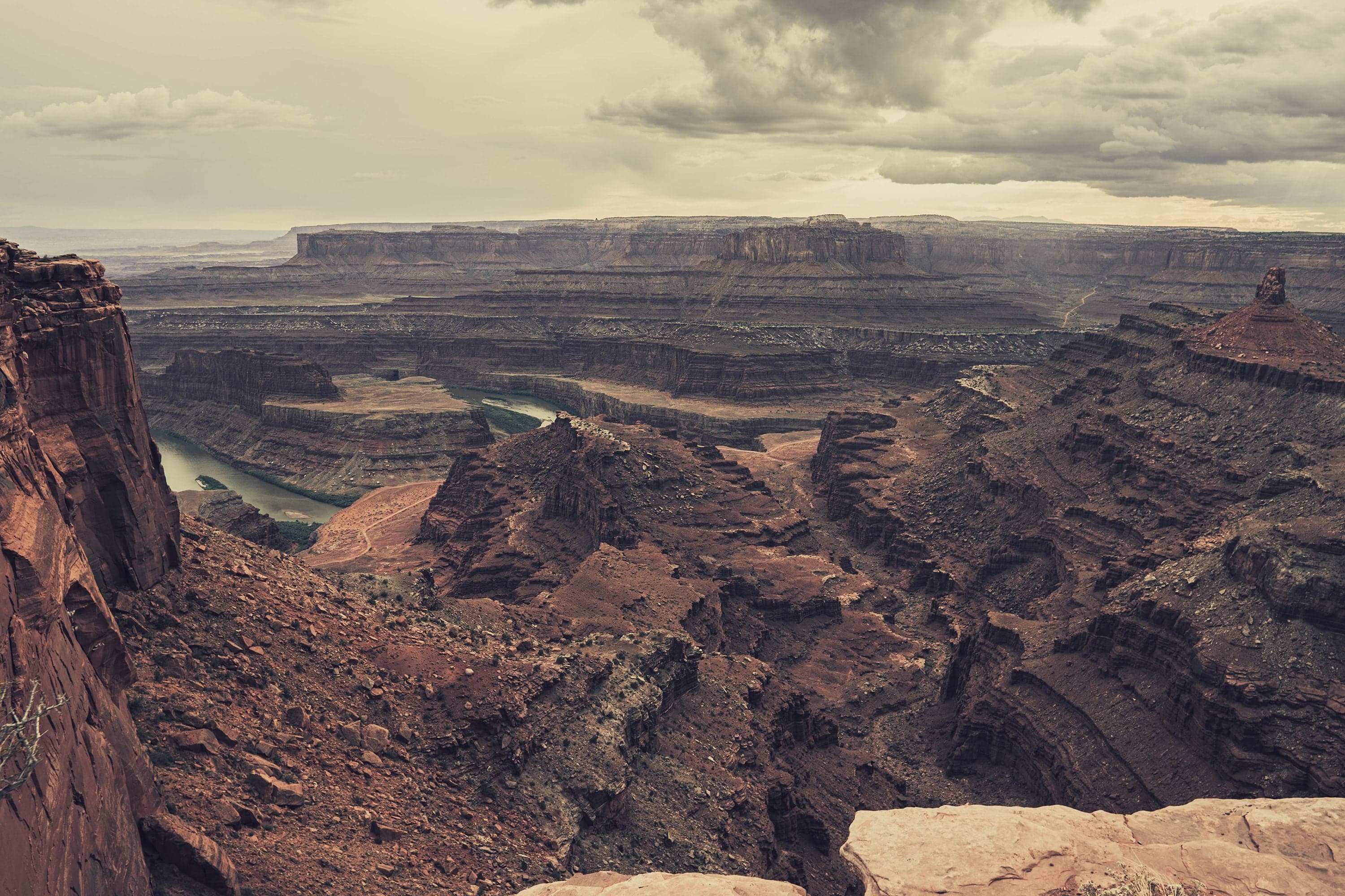 Storm clouds unleash visible rain shafts over the layered red-rock canyon at Dead Horse Point, golden light at the horizon.