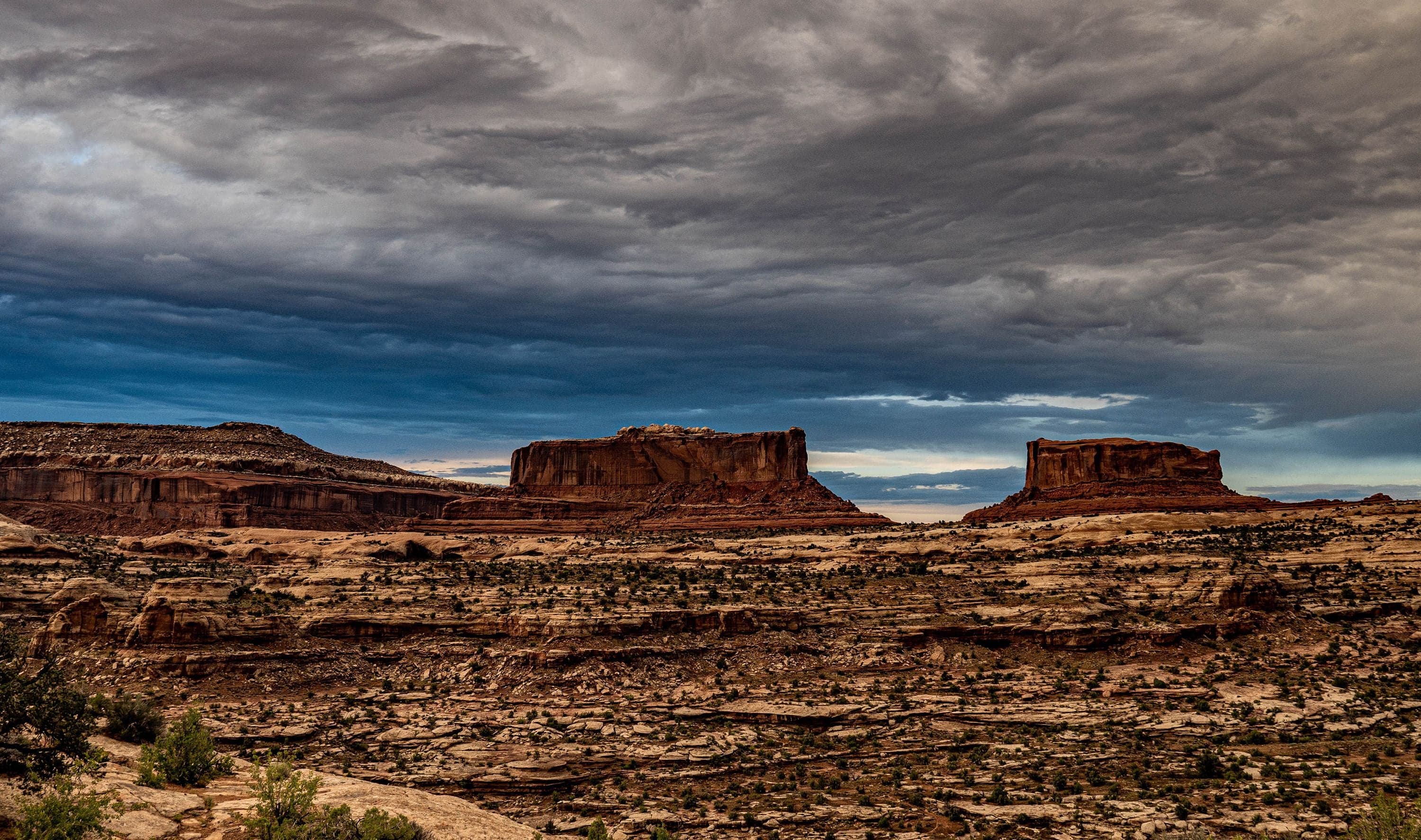 Two flat-topped red sandstone mesas rise from a rocky desert floor beneath a turbulent storm sky.