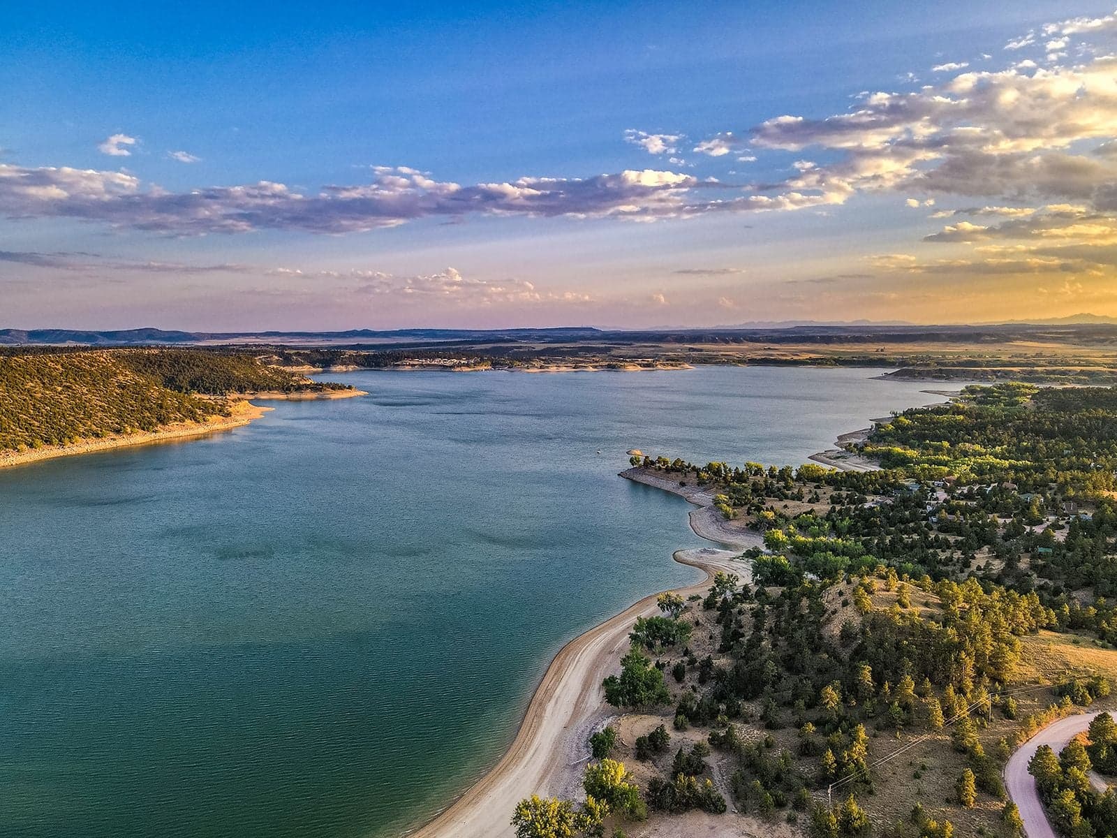 An aerial of a large Wyoming reservoir at sunset with sandy shoreline, pine-dotted banks, and a pastel blue-pink sky.