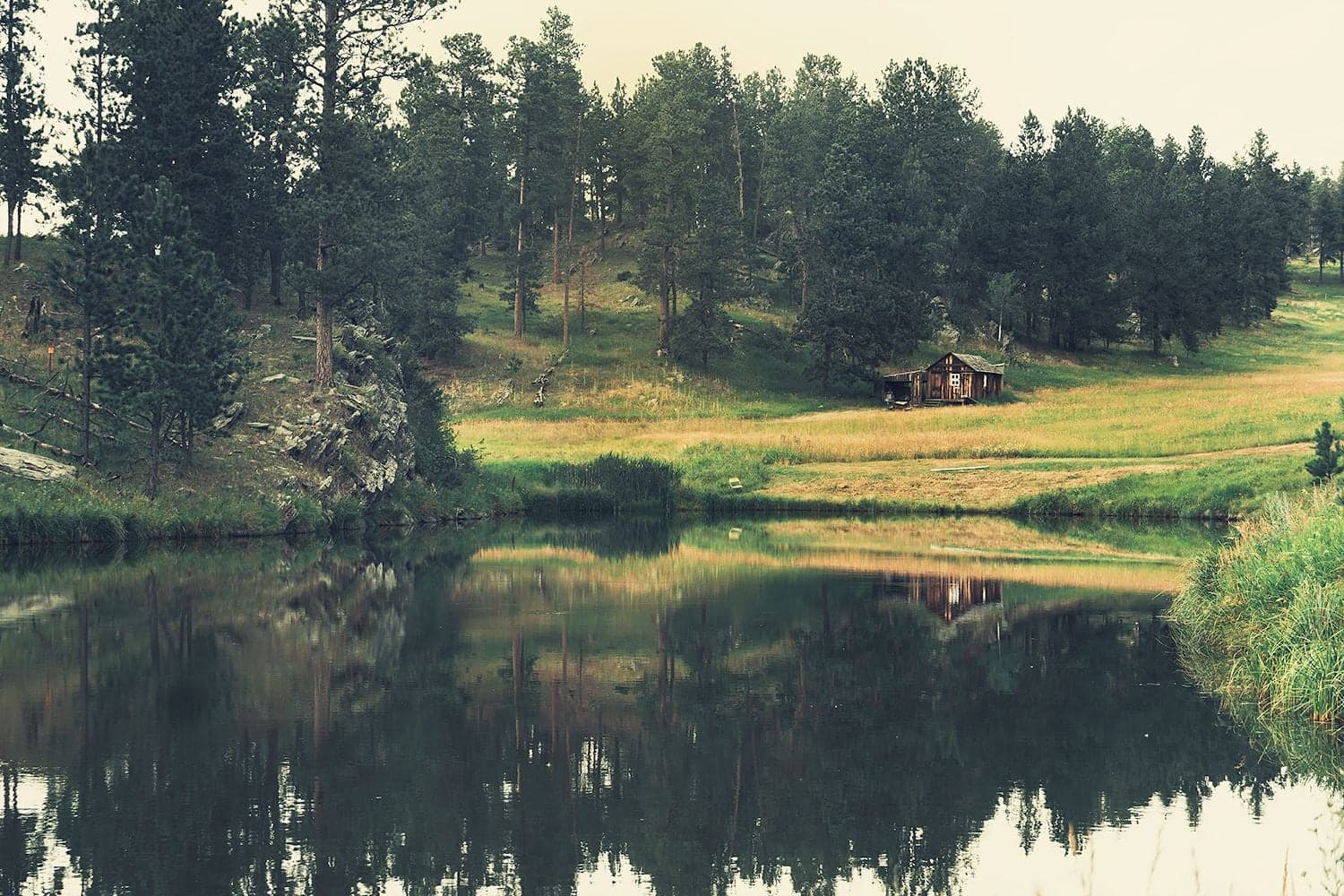 A small weathered wooden cabin sits on a grassy hillside above a glassy pond that mirrors the trees in muted tones.