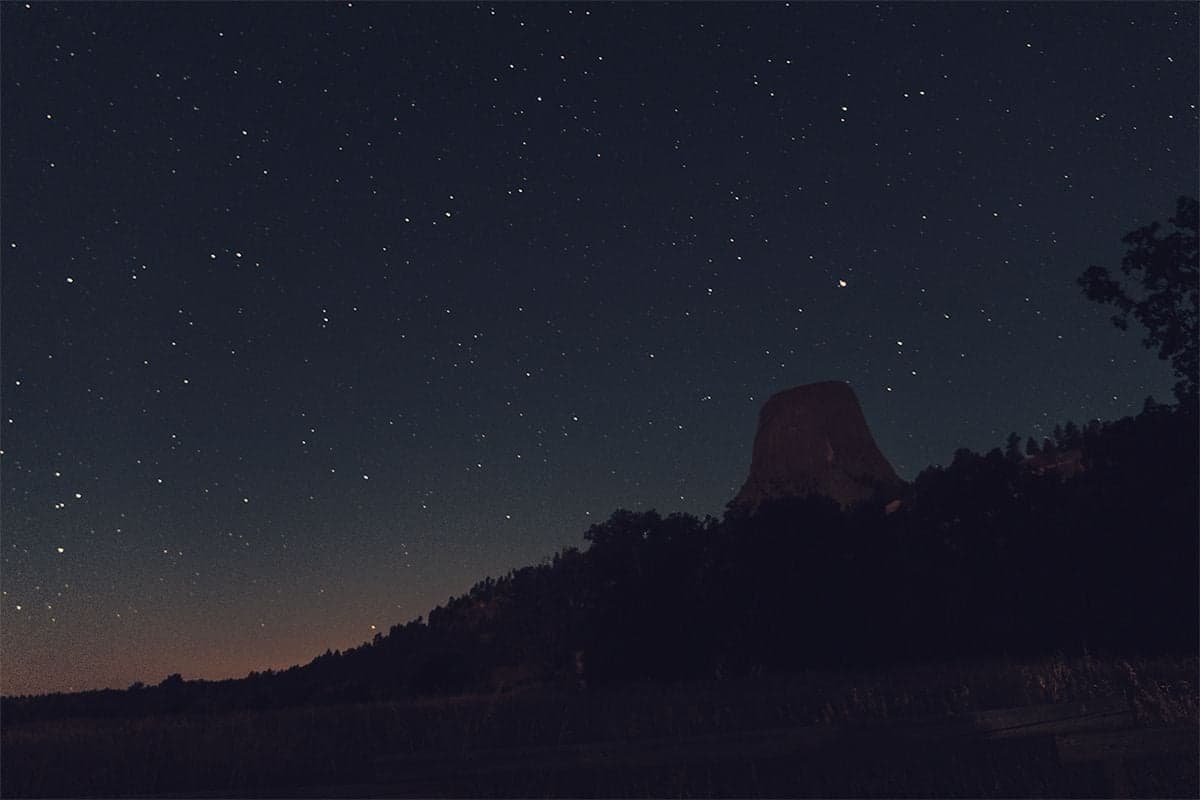 Devils Tower rises as a dark silhouette against a deep star-scattered sky with a faint warm glow on the horizon.