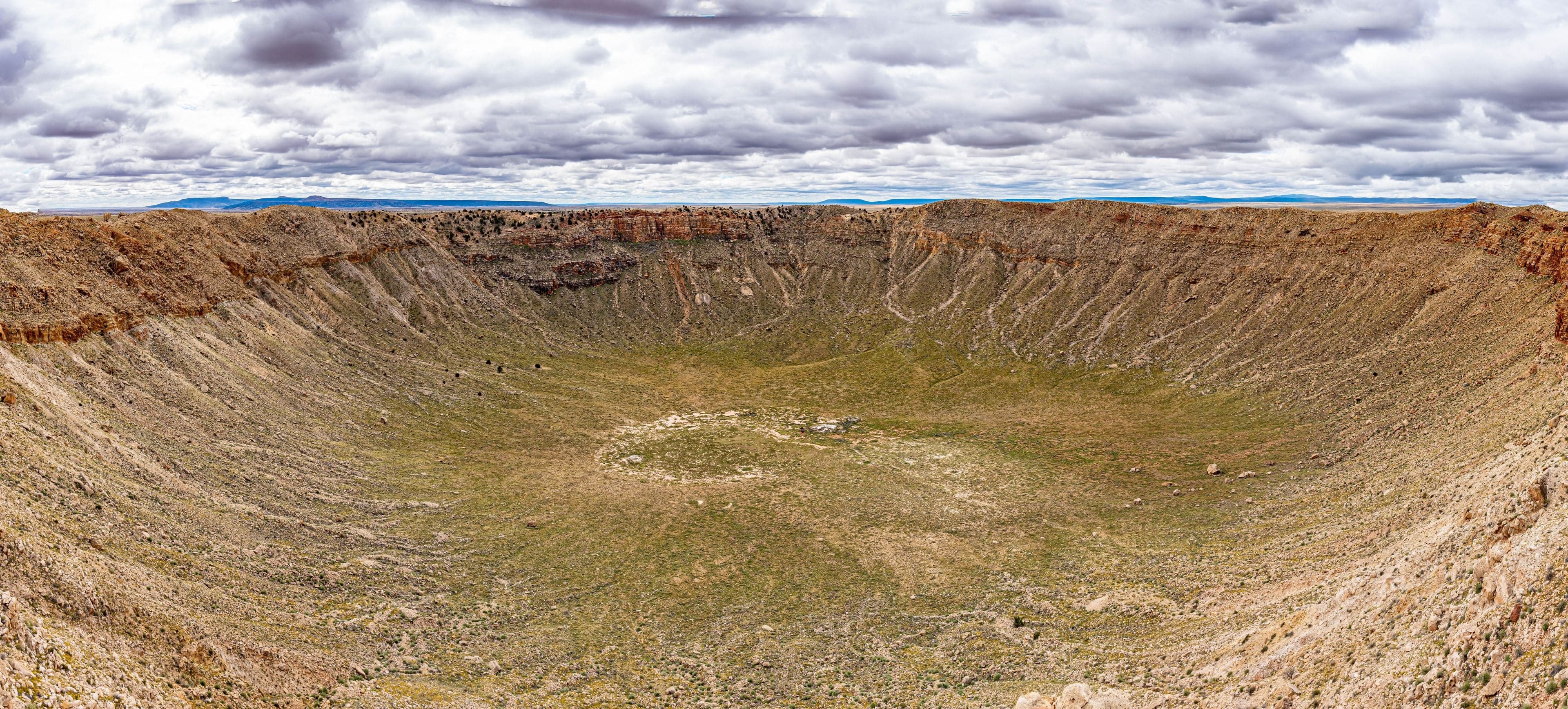 A wide panorama into the enormous bowl of Meteor Crater, layered rocky walls descending to a flat floor under heavy overcast.