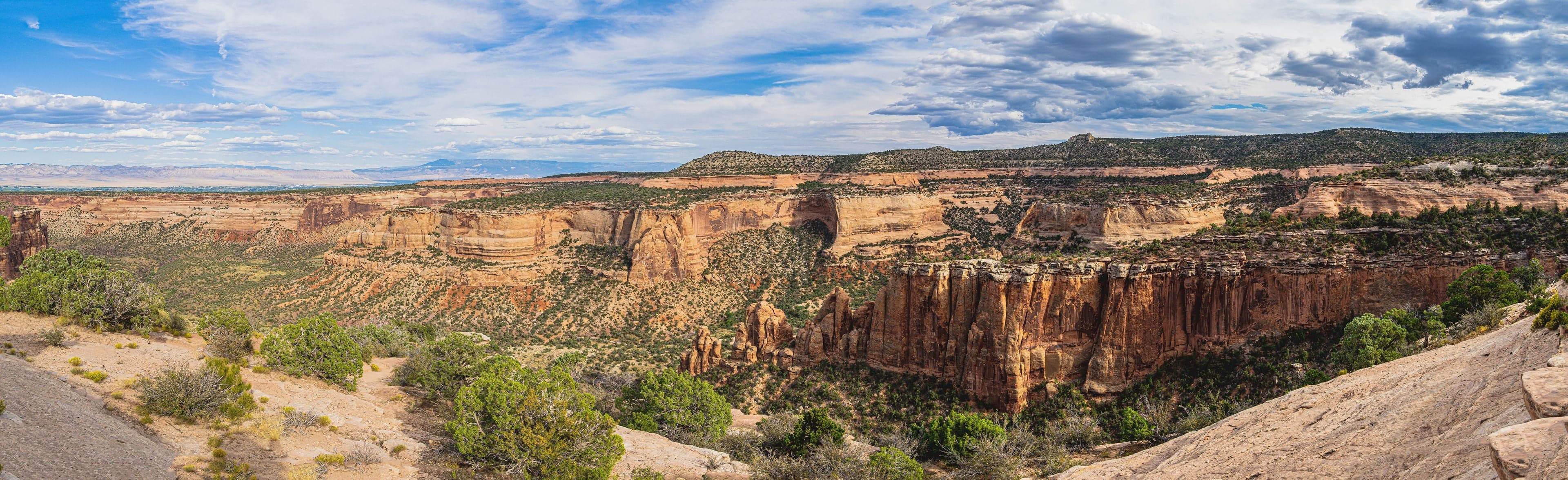 A panoramic overlook of sweeping red canyon cliffs and alcoves with a natural arch in the distance under rolling cumulus.