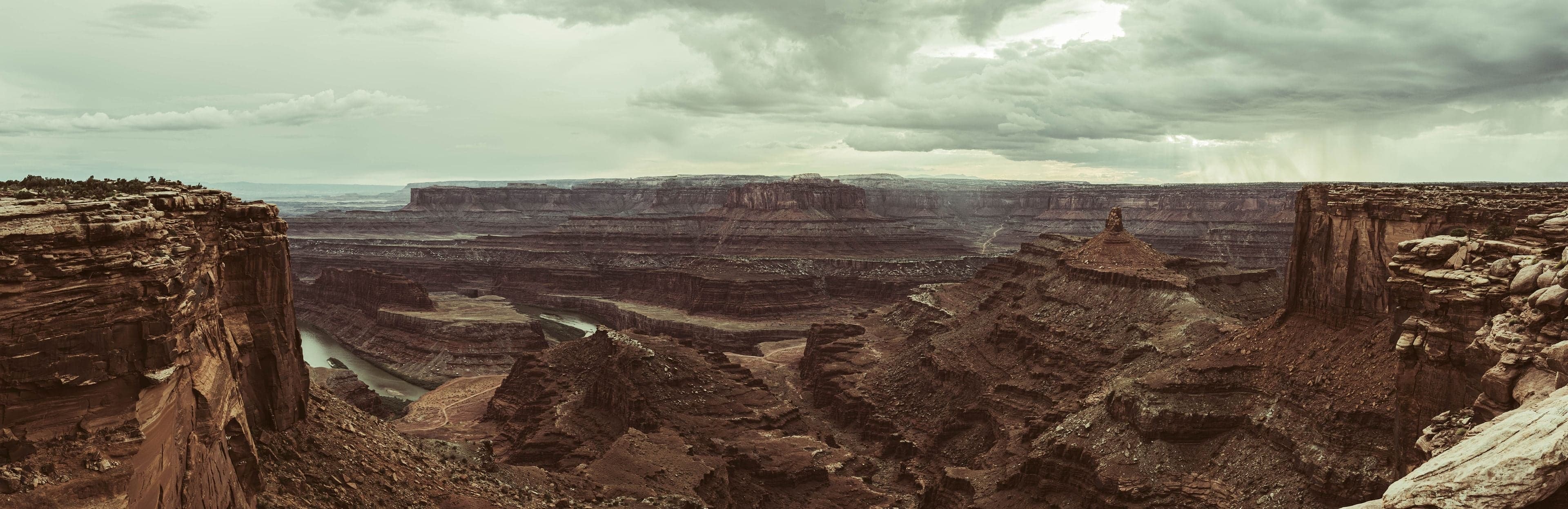 A panorama of the Dead Horse Point canyon and river gooseneck under stormy sky with blue light breaking at the right.