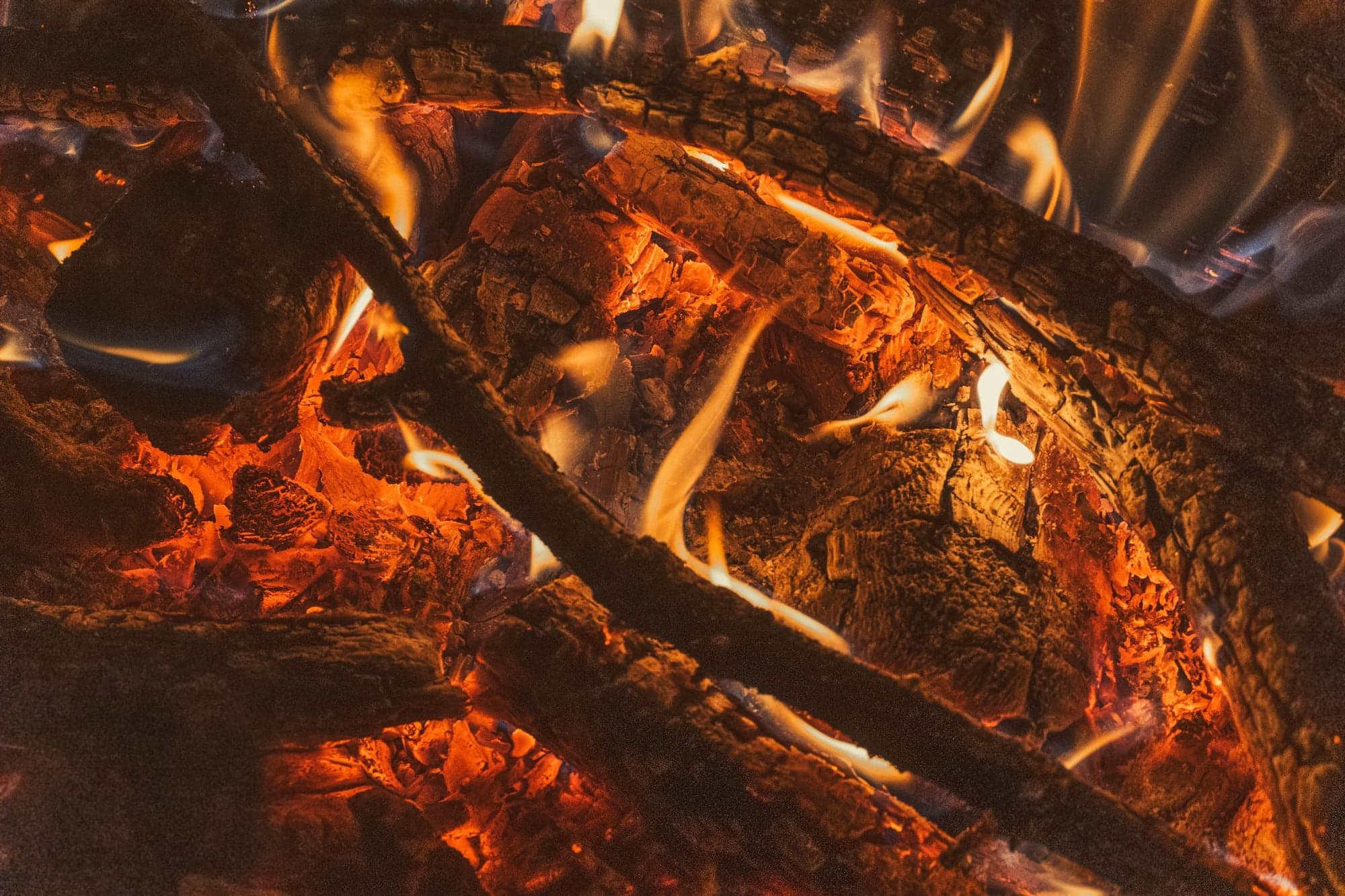 A close-up of campfire logs showing deep red-orange glowing embers and wispy flames against a dark background.