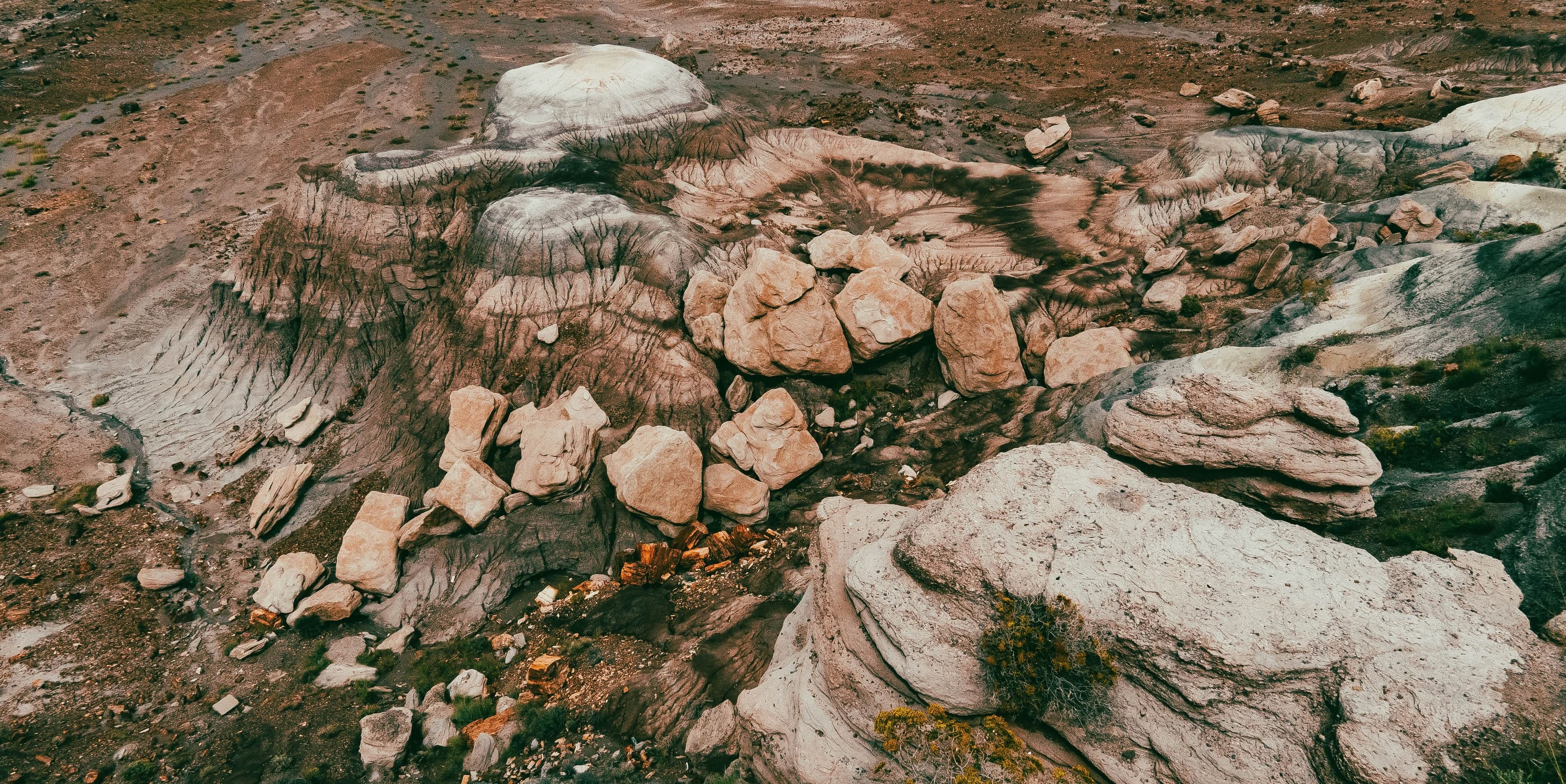 An aerial of an eroded badlands butte with a white caprock dome, dark streaked flanks, and scattered orange boulders.