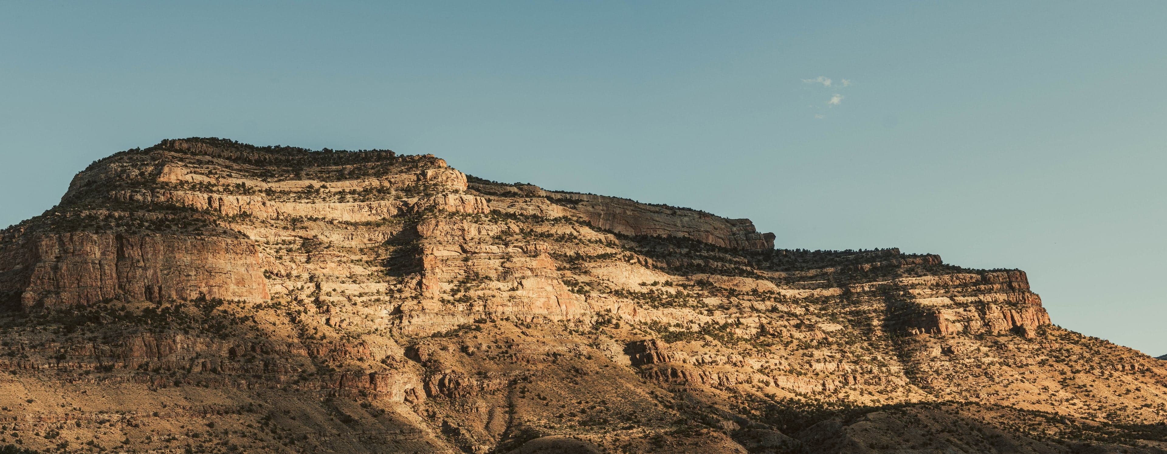 Late golden light illuminates the warm tan and rust layers of a broad sandstone mesa under a clear pale sky at dusk.