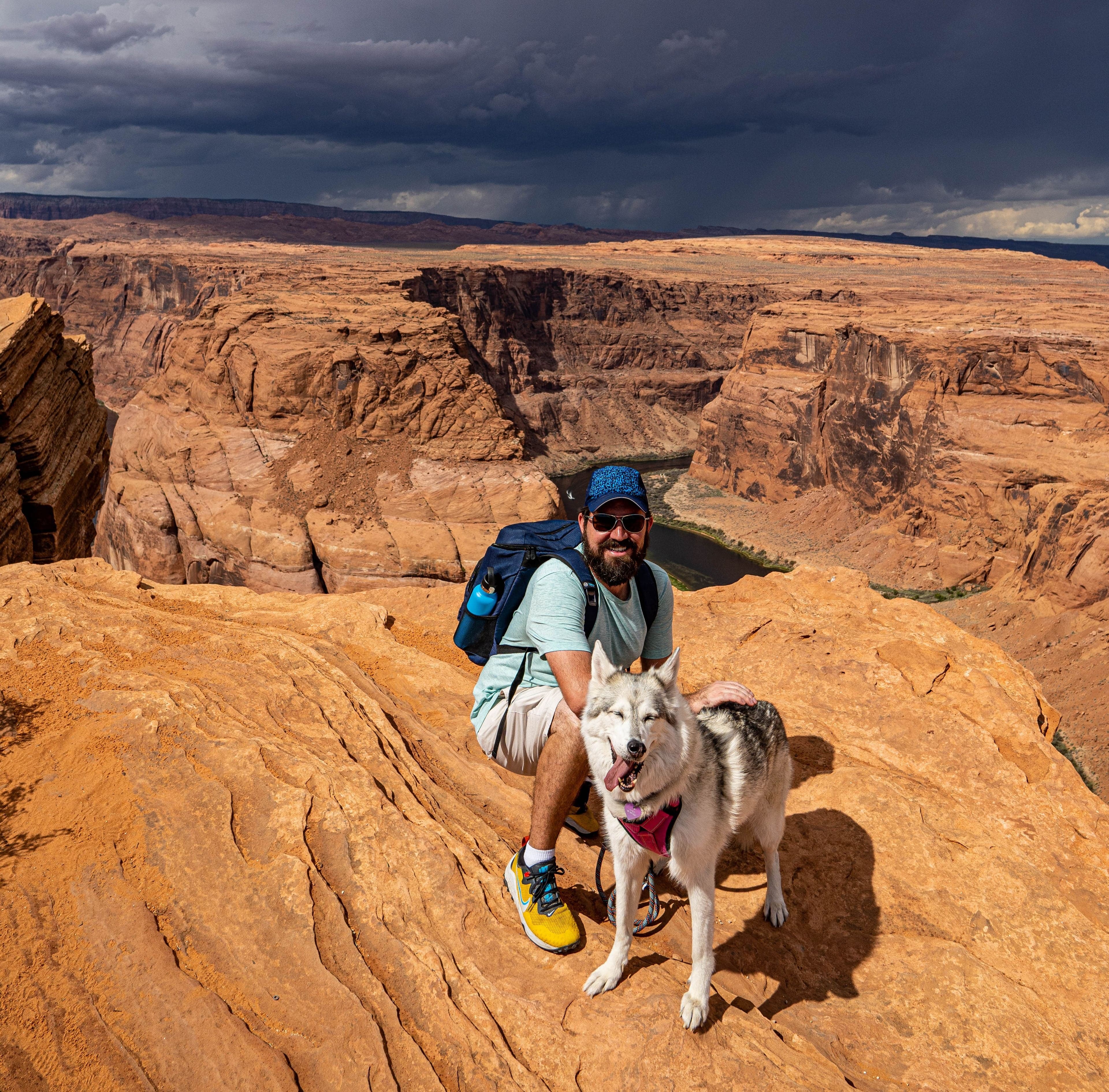A bearded man in sunglasses crouches beside a gray wolfdog on orange sandstone at Horseshoe Bend with the canyon and storm clouds behind.