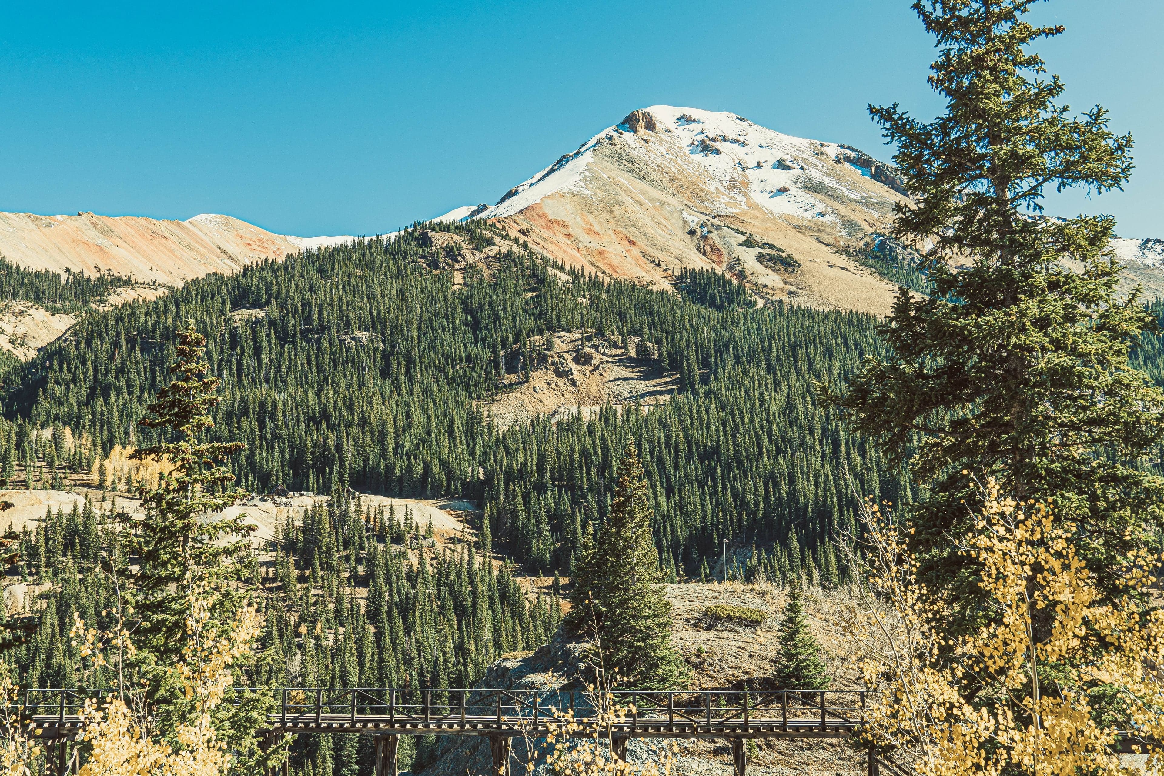 A snow-dusted peak rises above a valley of pine and golden aspen with a historic wooden trestle bridge visible through the foliage.