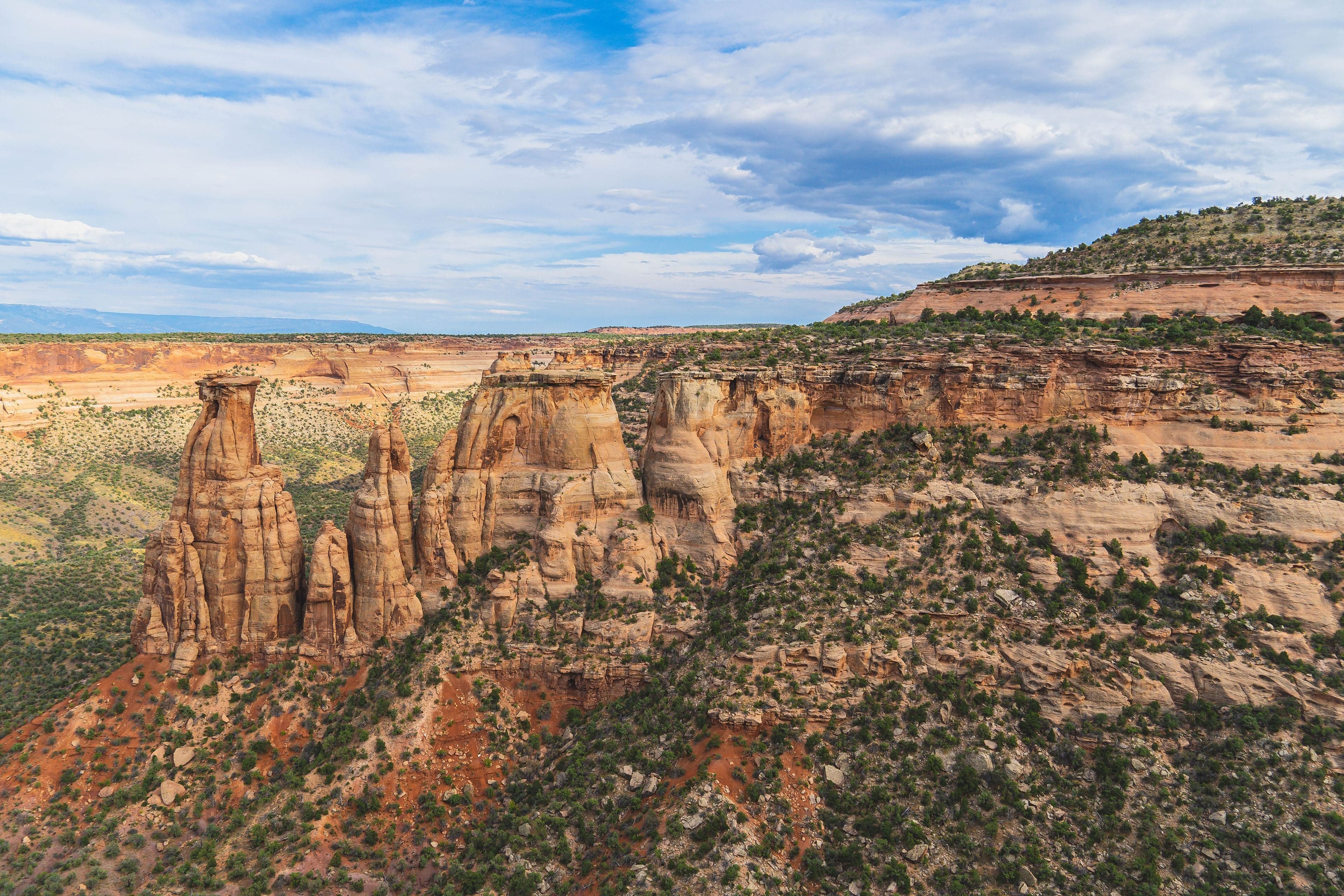 Three towering sandstone monoliths stand above a scrub-covered slope with a vast orange canyon and clouded sky behind.