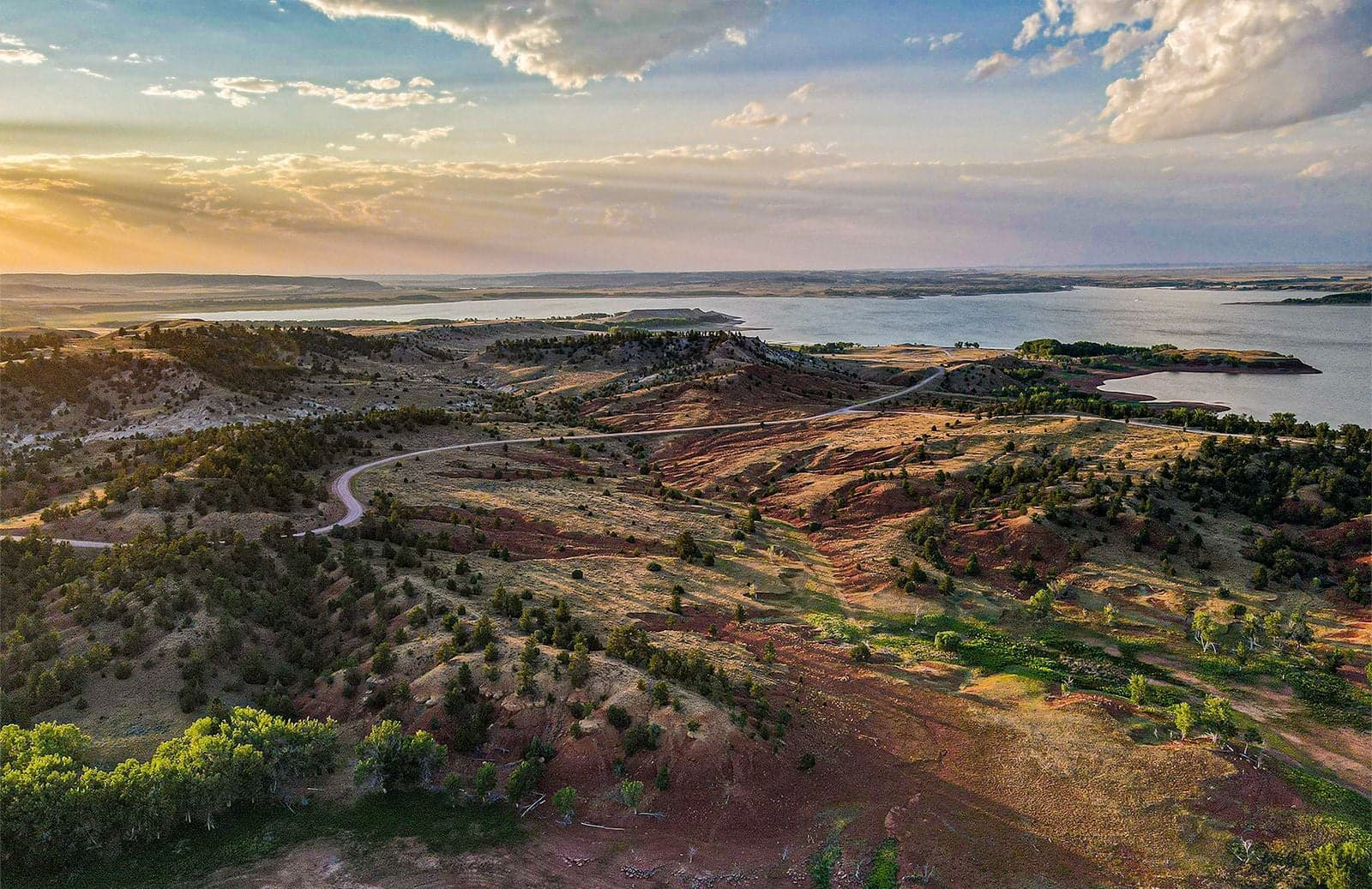 An aerial at sunset over rolling reddish Wyoming hills with a winding gravel road, scattered pines, and a reservoir beyond.
