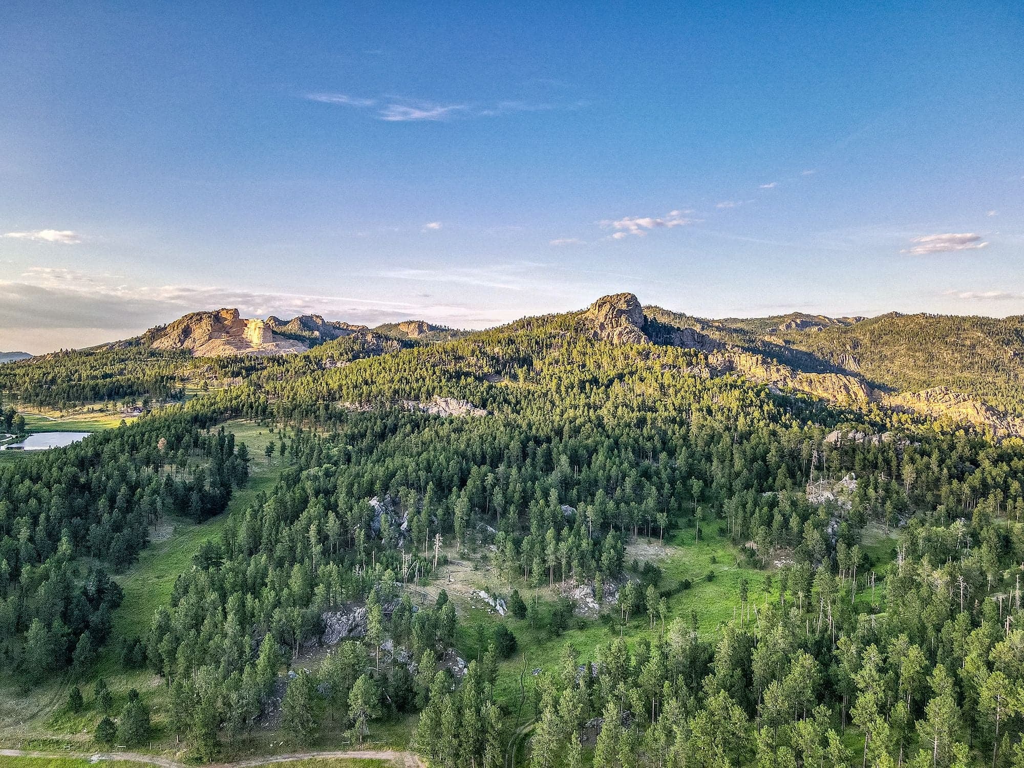 A sweeping aerial over vast green pine forest and rocky ridgeline at golden hour, the Crazy Horse carving faint on the skyline.
