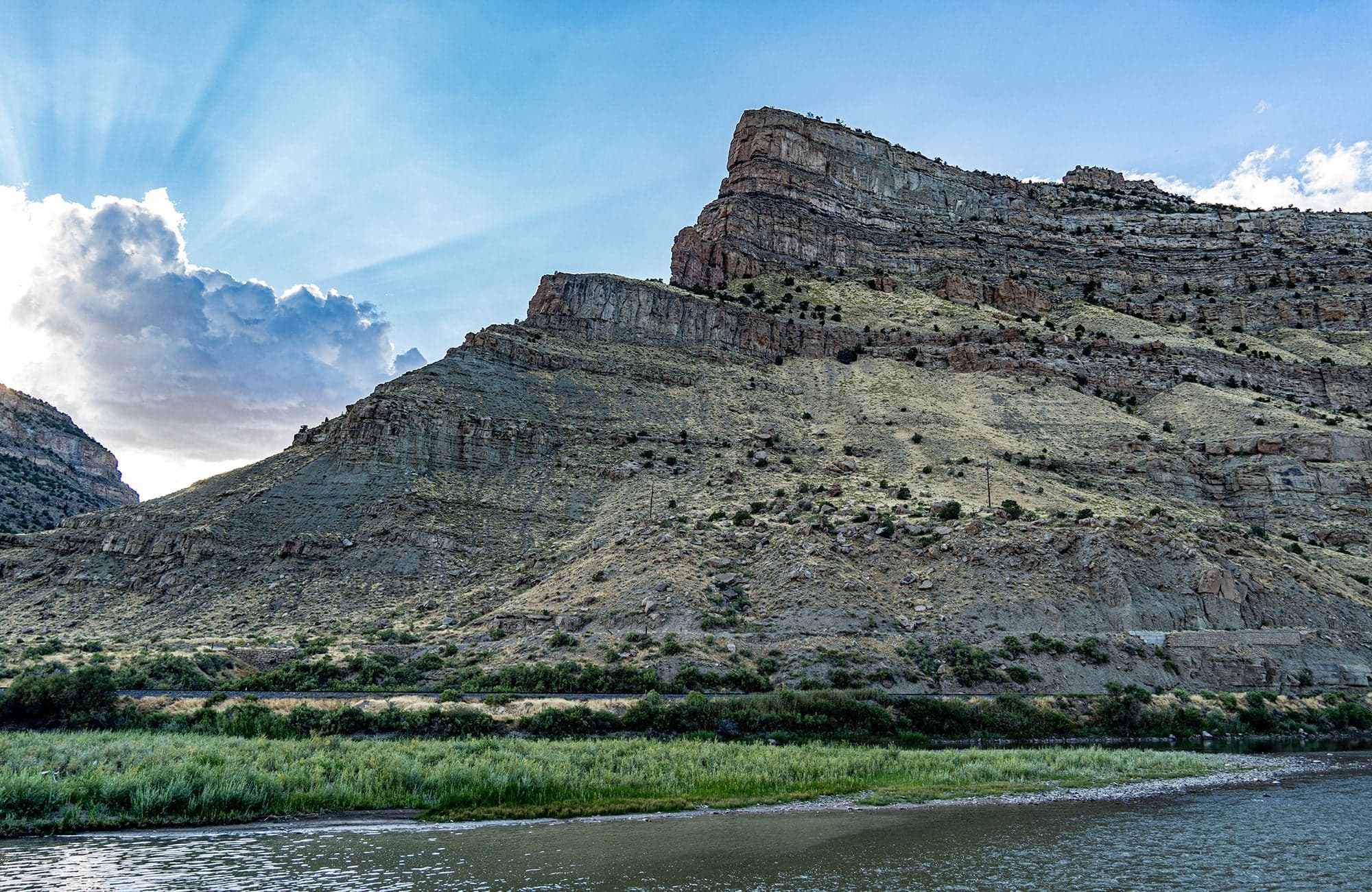The Colorado River flows along a grassy bank before a massive layered sandstone cliff, crepuscular rays fanning above.