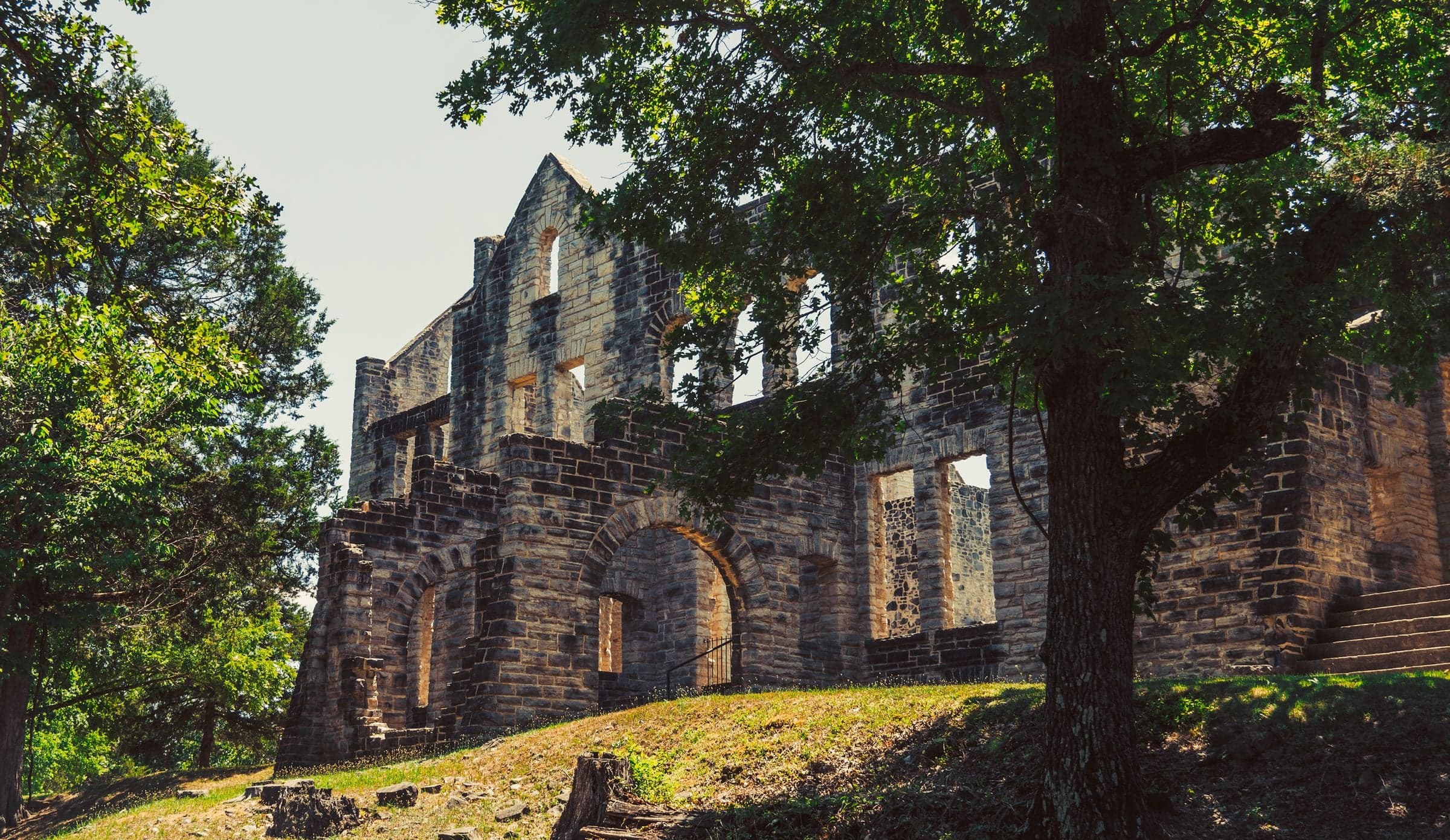 A roofless multi-story stone castle ruin sits on a grassy hillside surrounded by green oak and pine under pale overcast.