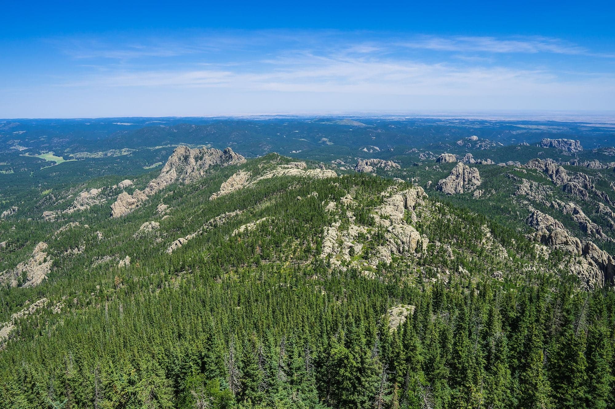 An aerial along a forested granite ridgeline toward distant plains, pine slopes falling away on both sides.