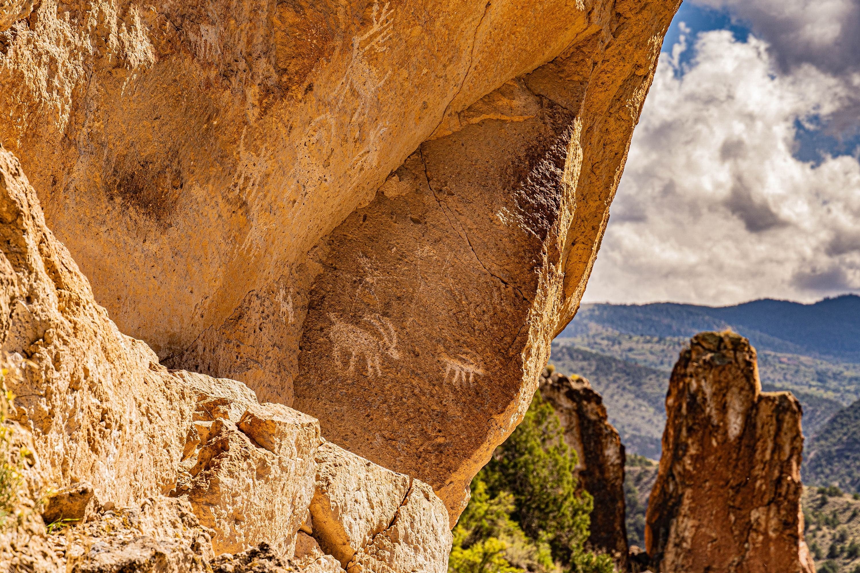 Fremont petroglyphs of a deer and handprint on a sandstone overhang, framed by a rock alcove opening onto a mountain valley.