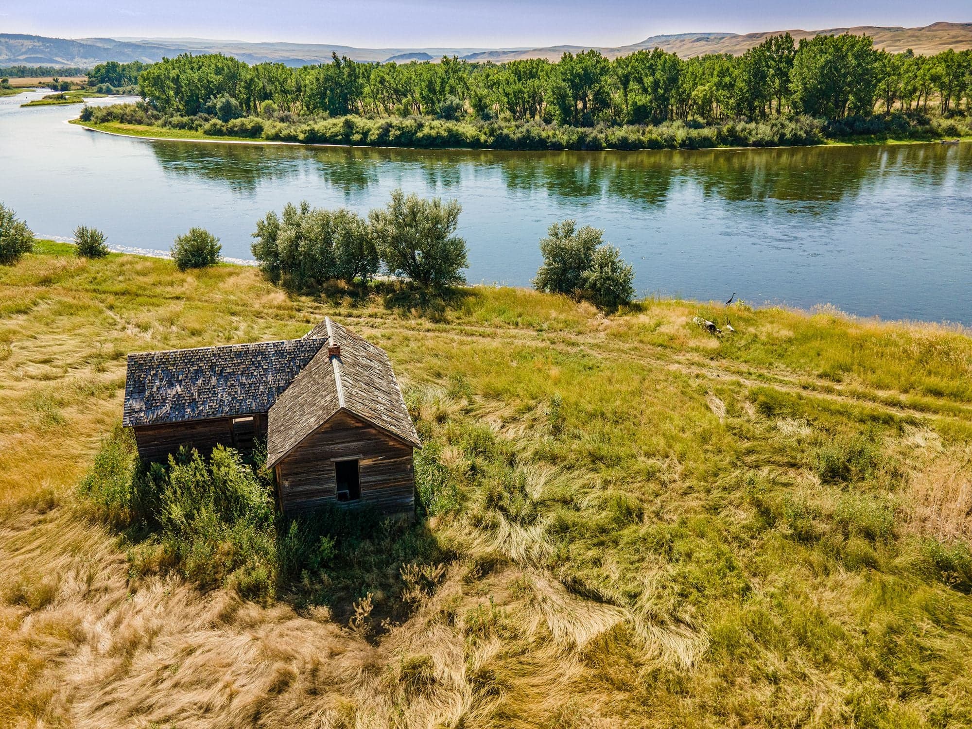 An aerial of a small deteriorating wooden shack in golden dry grass on the bank of a wide meandering cottonwood-lined river.