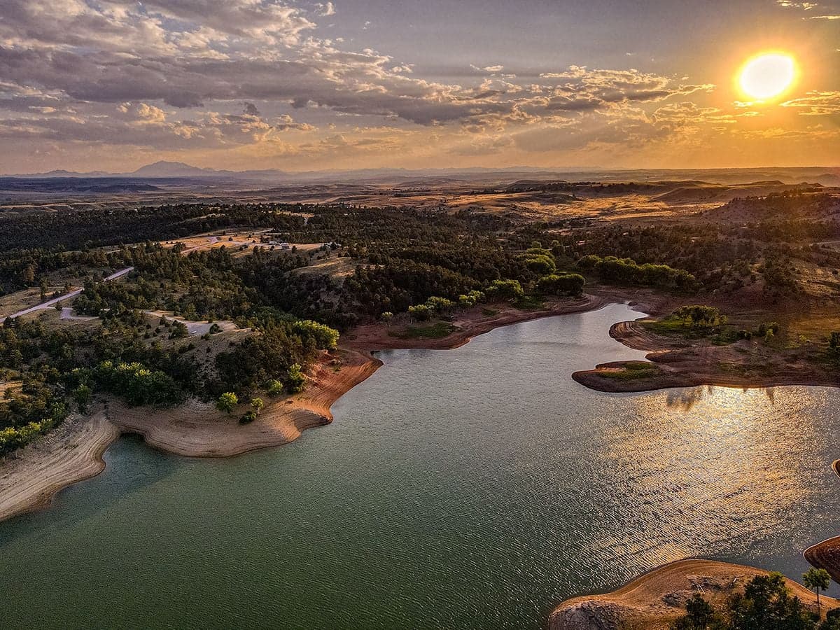 An aerial of a winding lake shoreline at sunset with golden light on the water, green trees, and a warm glowing sky.