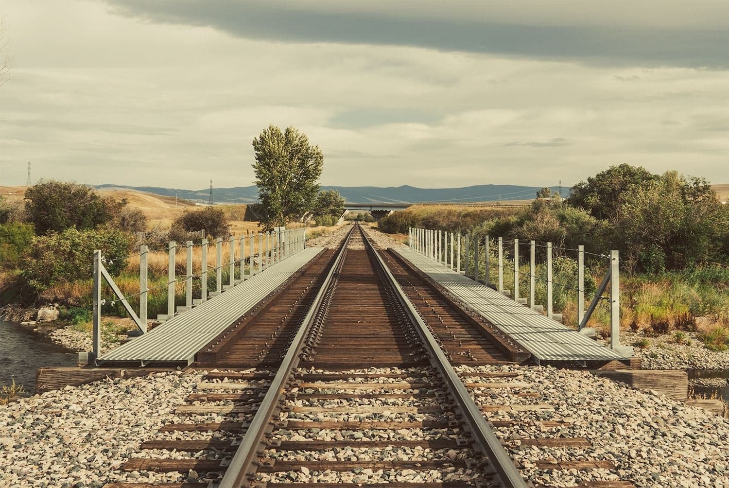 Railroad tracks on a flat bridge with metal guardrails converge toward a vanishing point across wide Western grassland under overcast.