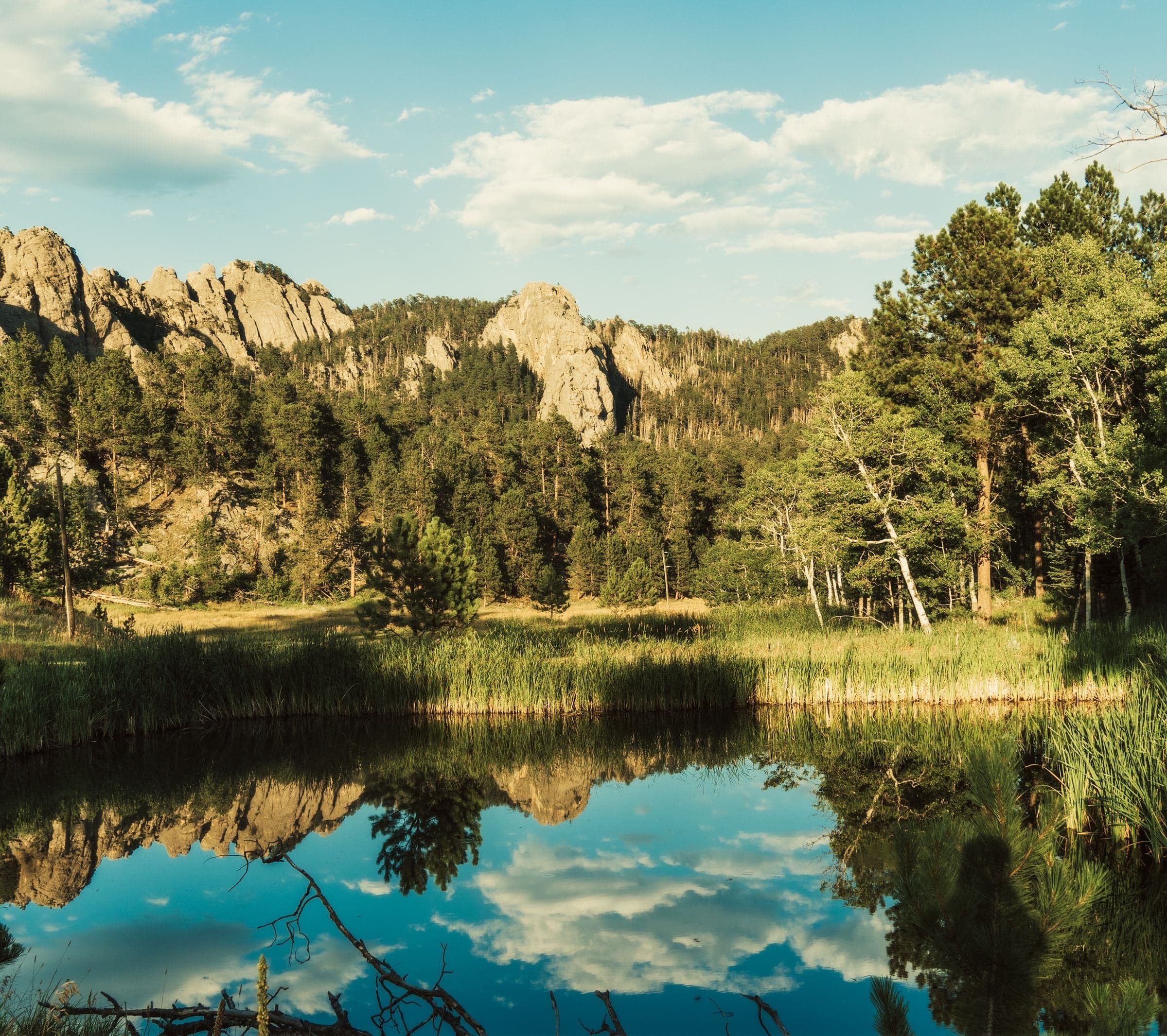 A tranquil pond reflects towering granite formations and a pine hillside under a partly cloudy sky in warm late light.