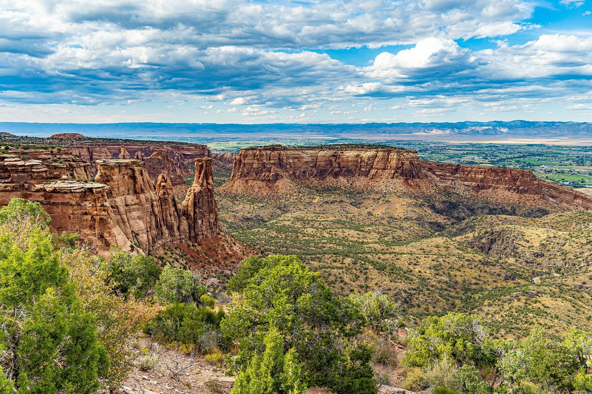 A slender sandstone spire rises from a desert bench before a flat-topped mesa, the green valley and distant mountains beyond.
