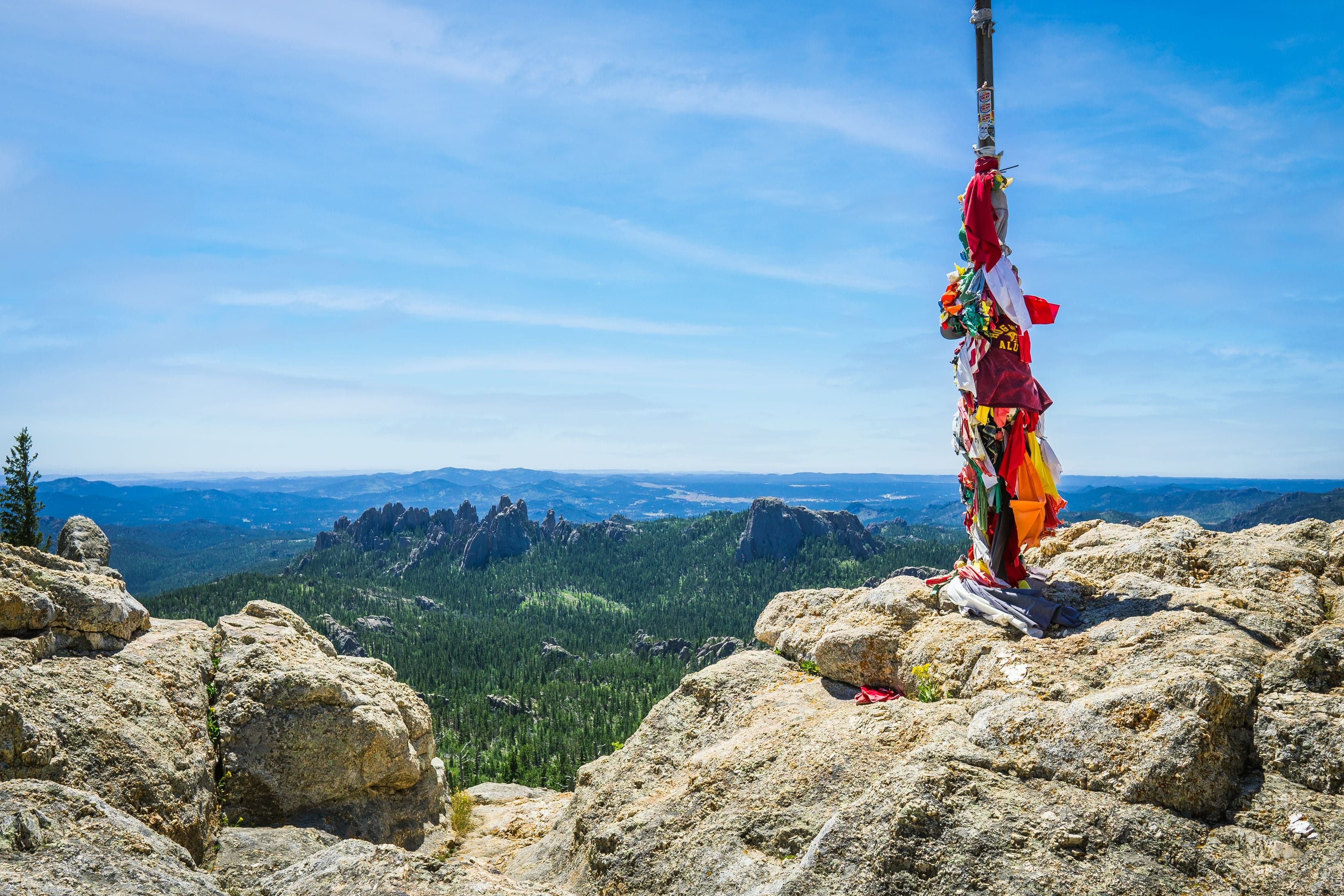 A pole covered in colorful tied fabric offerings stands on the granite summit of Black Elk Peak above a sweeping Black Hills panorama.