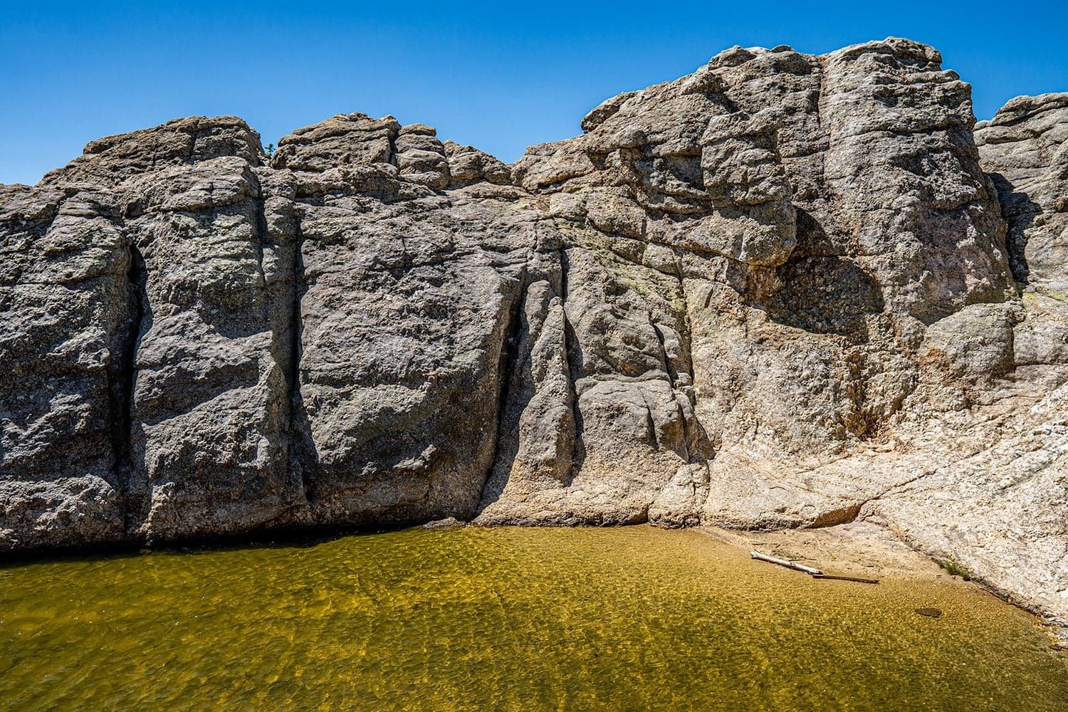 Tall weathered gray limestone formations rise above a shallow green-tinted pool against a clear blue sky, driftwood on the shore.