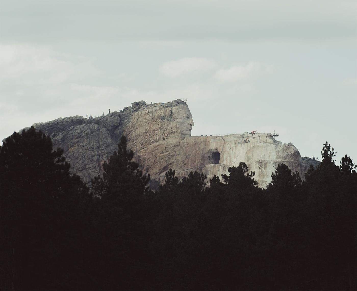 The partially completed Crazy Horse Memorial carving juts from a granite mountain, framed by dark conifer silhouettes against grey sky.